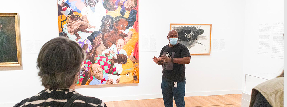 A man gestures toward a painting in a museum while a group looks on.