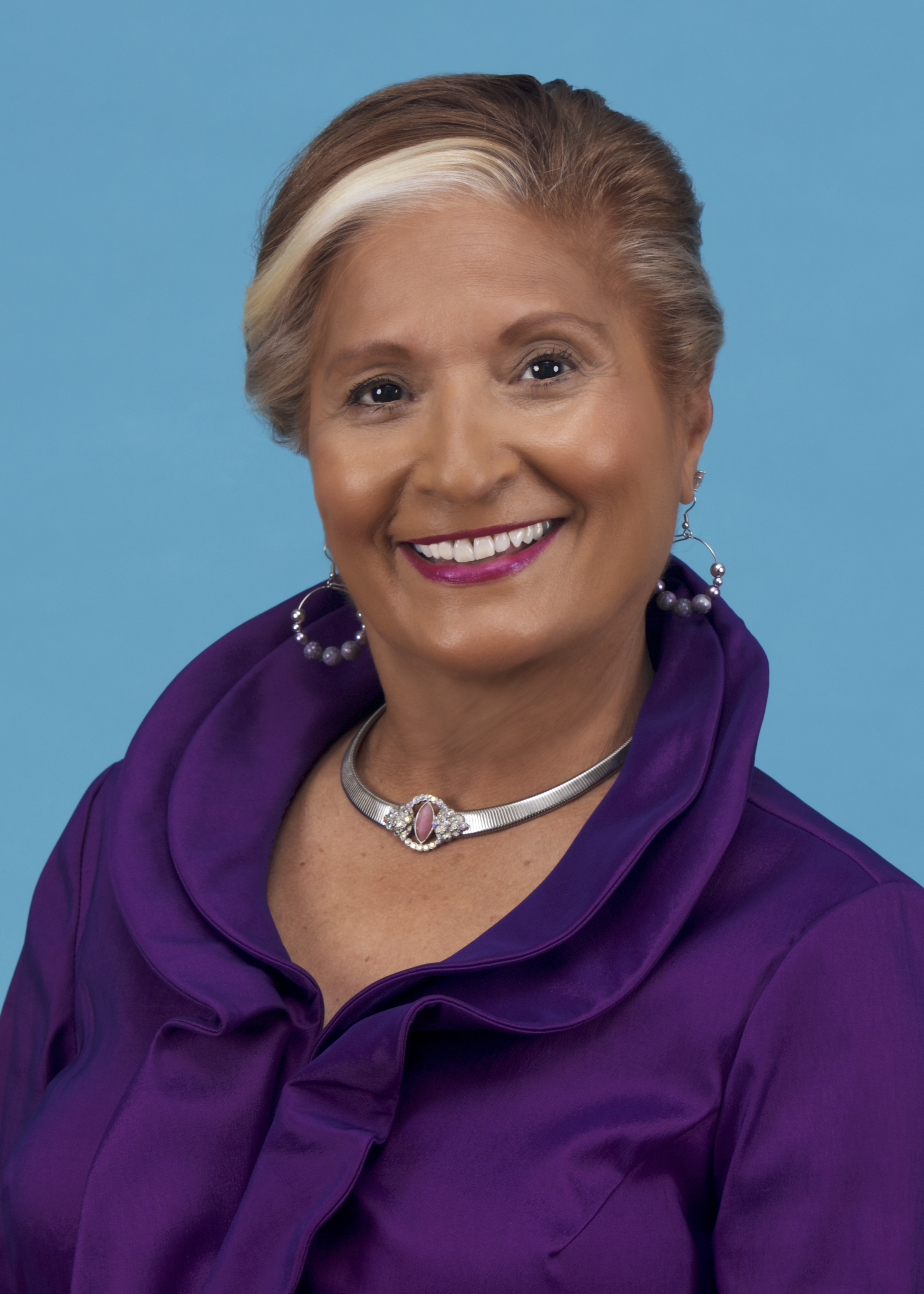 Latina women with highlights pulled up smiling and wearing a purple blouse and necklace.