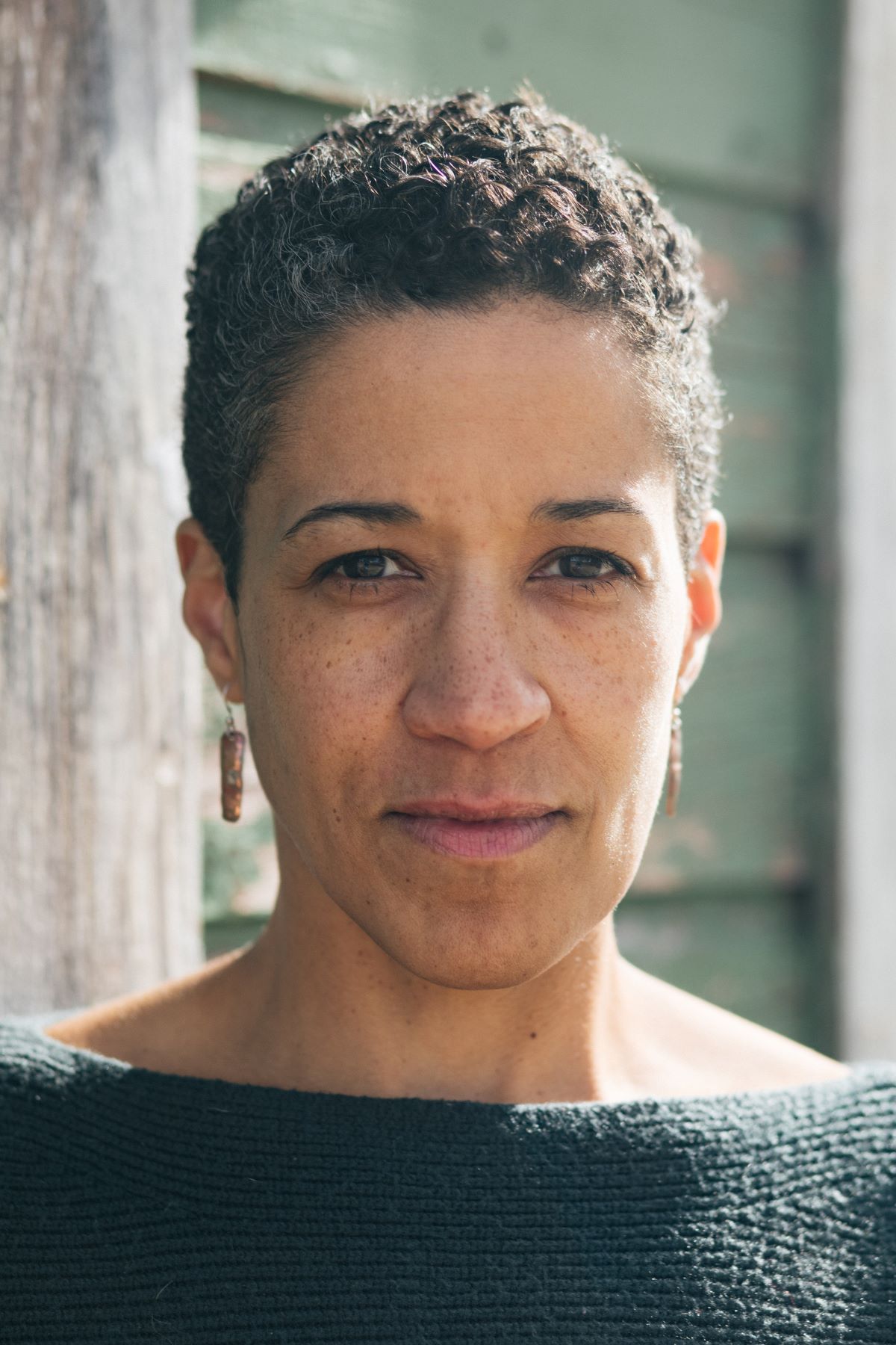 Woman with short, curly hair, wearing earrings and a textured dark top. She has light brown skin with subtle freckles, dark eyes, and a neutral expression.